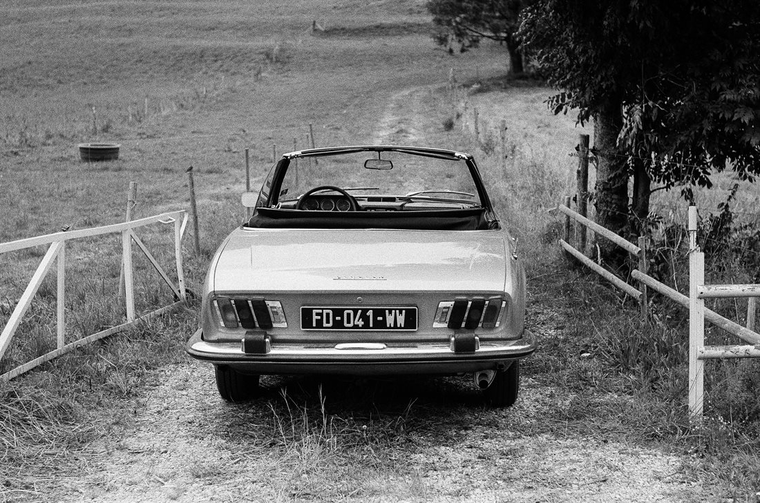 Cantal, noir et blanc, voiture Peugeot 504 Cabriolet de collection vue de l'arrière, capote baissée, à l'entrée d'un pré barrière bois ouverte